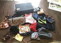 Multiple purses and handbags arranged on a wooden floor, showing variety in materials and colors, plus a black storage drawer.