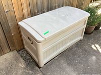 Suncast outdoor storage bin placed against wooden fence, showing front and side.
