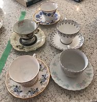 Group photo of five different vintage teacups and saucers on a speckled counter, showing various floral and decorative patterns.