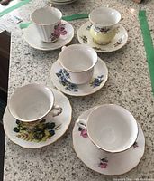 Five assorted vintage teacups with matching saucers on a stone countertop, showing different floral designs and gold rims.