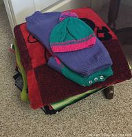 Stack of various beach and pool towels with a knitted kids hat and sweater set on top, showing colors and details of the items.