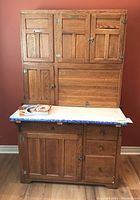 Front view of cabinet showing top hutch, white laminate countertop with blue edge trim, bottom storage with door and drawers, and two cookbooks on countertop.