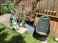 Photo showing small black and green plastic wheelbarrow, garden hose on reel, three snow shovels with red grips, two green tomato cages, garden fork tool, and a bag of soil/fertilizer.