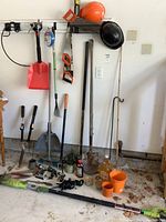 Photo showing a collection of garden hand tools including shovels, rakes, pruners, safety helmets, a red dustpan, green hose, and the electric pole saw lying on the floor
