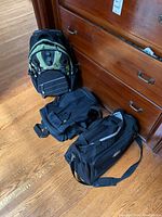 Photo showing the three bags together: a green and black backpack, and two black duffle bags.