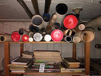 Shelf containing stacked books including cooking and art books, and tubes with rolled posters having red and black lids.
