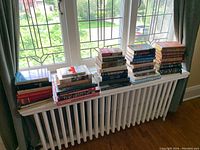Wide view showing stacks of books on a radiator under a window, showcasing a variety of sizes and thicknesses indicating assorted genres and authors.