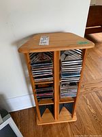 Wooden CD rack holding multiple CDs in jewel cases, shown from slightly angled front view on wooden floor against a white wall.
