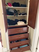 Wardrobe with folded assorted men's and women's clothing on shelves and in drawers. Clothing includes dark pants, jeans, and patterned fabric items.