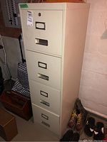 Full view of vertical metal filing cabinet with four drawers, off-white color, displaying wear and some scuffs. Cabinet located in basement setting.