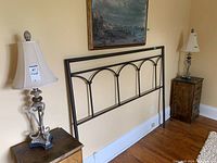Two matching table lamps with metal bases and beige fabric shades placed on wooden side tables next to a metal bed headboard.