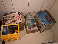 Four boxes of vintage magazines on floor by wall, showing various titles and stacks.