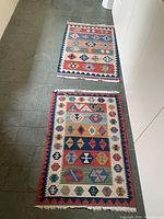 Two rectangular cotton mats on floor, each with colorful geometric patterns and fringed edges, showing full design and scale