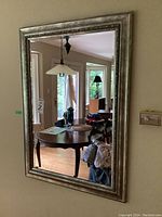 Bevelled rectangular mirror hung on wall showing wooden floor, window, and furniture reflected in it.