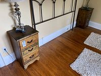 Both vintage wooden side tables positioned on hardwood floor with decorative headboard behind. Each has three drawers, metal handles, and visible scuffs and scratches.