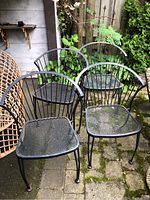 Group photo of four black metal outdoor chairs with mesh seats and curved backs on an outdoor paved area near plants.