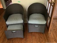 Front view of two brown resin wicker chairs with light grey seat cushions and footstools partially extended on hardwood floor against a wall.
