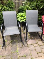 Two black metal framed rocking chairs with gray mesh fabric seats placed outdoors on patio stones with greenery and red Adirondack chair in background.