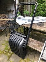 First photo shows one black snow scoop shovel with deep curved scoop and metal tubular handle leaning against wooden table in outdoor patio.