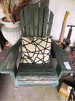 Photo of a green wooden Adirondack chair with wide armrests and a beige pillow with black geometric lines in an indoor garage setting. Chair shows signs of wear and chipped paint.