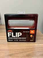 JBL Flip portable wireless speaker in original box packaging showing speaker through clear window with branding and features on box front.