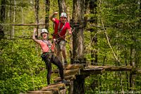 Two people enjoying an aerial trek on a wooden suspended bridge wearing helmets and harnesses in a forested area.