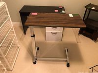 Full view of the hospital bed table with wood grain rectangular top, metallic adjustable height frame, and four caster wheels for mobility.