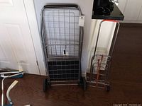 Two wire folding carts: a large grocery cart with black wheels and a smaller luggage cart with red handle, standing on a wooden floor against a white wall.