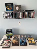 Metal wall-mounted CD shelf with CDs and DVDs arranged on and below it. Visible titles include The Lord of the Rings boxed set, various albums of music and films.