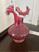 Photo showing the front view of the cranberry glass vase with white glass trim around the fluted top edge and the swirled texture on the body.