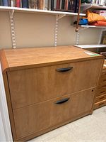View of two-drawer lateral filing cabinet showing wood finish and black handles, placed against wall under shelves.