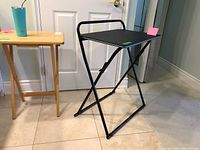 Wood TV tables and black folding table shown from an angle in a room with tile floor. Two wooden tables with natural finish and a black metal frame folding table.