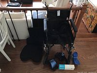 Photo showing black folding wheelchair with removable pedals and memory foam cushions placed beside and on it, plus blue air cast foot braces and black and blue hip and knee braces lying on wooden floor.
