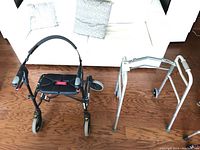 Two walkers side by side: black rollator with four wheels and silver folding walker
