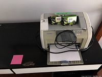 Front view of HP LaserJet 1020 printer showing extra HP 933 XL color cartridges on top, USB cable coiled on the printer, paper in the tray.