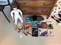 Collectible doll standing on a white stand with no clothing, in front of a wooden dresser. Four knitted hats and ten books laid out in front, including Bible, Black Beauty, and others. Two wooden crosses are placed near the doll's feet.