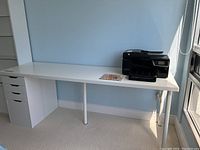 Full view of desk with a white tabletop, three detachable cylindrical white metal legs, and a white set of drawers featuring three smaller drawers and one large bottom drawer on the left side. Printer and desk mat on top not included.