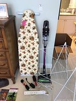 Photo showing the ironing board, wire drying rack, Bissell purple cordless vacuum, white extension cord, and power strip with other small accessories grouped on floor