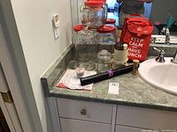 Photo showing five glass canisters with red lids, a glass jar with glass lid, electric coffee grinder, red insulated lunch bag, kitchen timer, paper doilies, cupcake foil liners, and black cooking mat on a bathroom countertop next to a sink.
