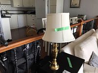 Brass table lamp with cream shade sitting on dark surface in living room near couch and kitchen in background.