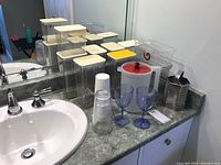 Overview of clear plastic kitchen canisters with lids, plastic cups, storage bins, wine glasses, jug, and grater on bathroom countertop.