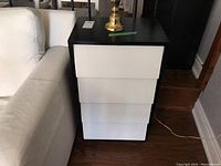 Black cabinet side table with four white drawers seen next to white couch. Shows overall shape and drawer fronts.