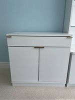 Front view of the white cupboard showing drawer and double door access with metal handles.