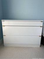 Front view of a white dresser with three drawers and gold handles on the right edge of each drawer.