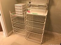 Photo showing two white stackable wire basket organizers with 3 and 4 wire baskets respectively, a small white plastic 4-drawer organizer on top, all on the floor.