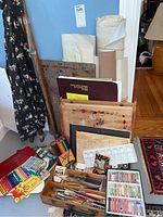 Photo showing easel, rolled canvas or paper, sketch pads, colored pencils, and sets of pastels and brushes on floor in corner.