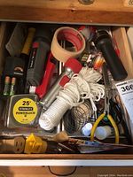 Top view of a partially open drawer containing household catch-all items such as measuring tape, batteries, string, flashlights, tape, wire cutters, and spray can.