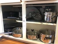 Wide view of kitchen cabinet containing several cookware items including stainless steel pots with glass lids, roasting pans, black grill pans, and cooking racks