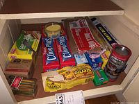 Shelf with various boxes of food wraps including Saran Cling Plus, yellow baking cups, Sterno cans, toothpicks, and sandwich bags.