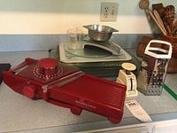 Photo showing red KitchenAid mandolin slicer, mechanical kitchen scale with round dial, glass cutting boards stacked, stainless steel mixing bowl and metal cheese grater on kitchen counter.